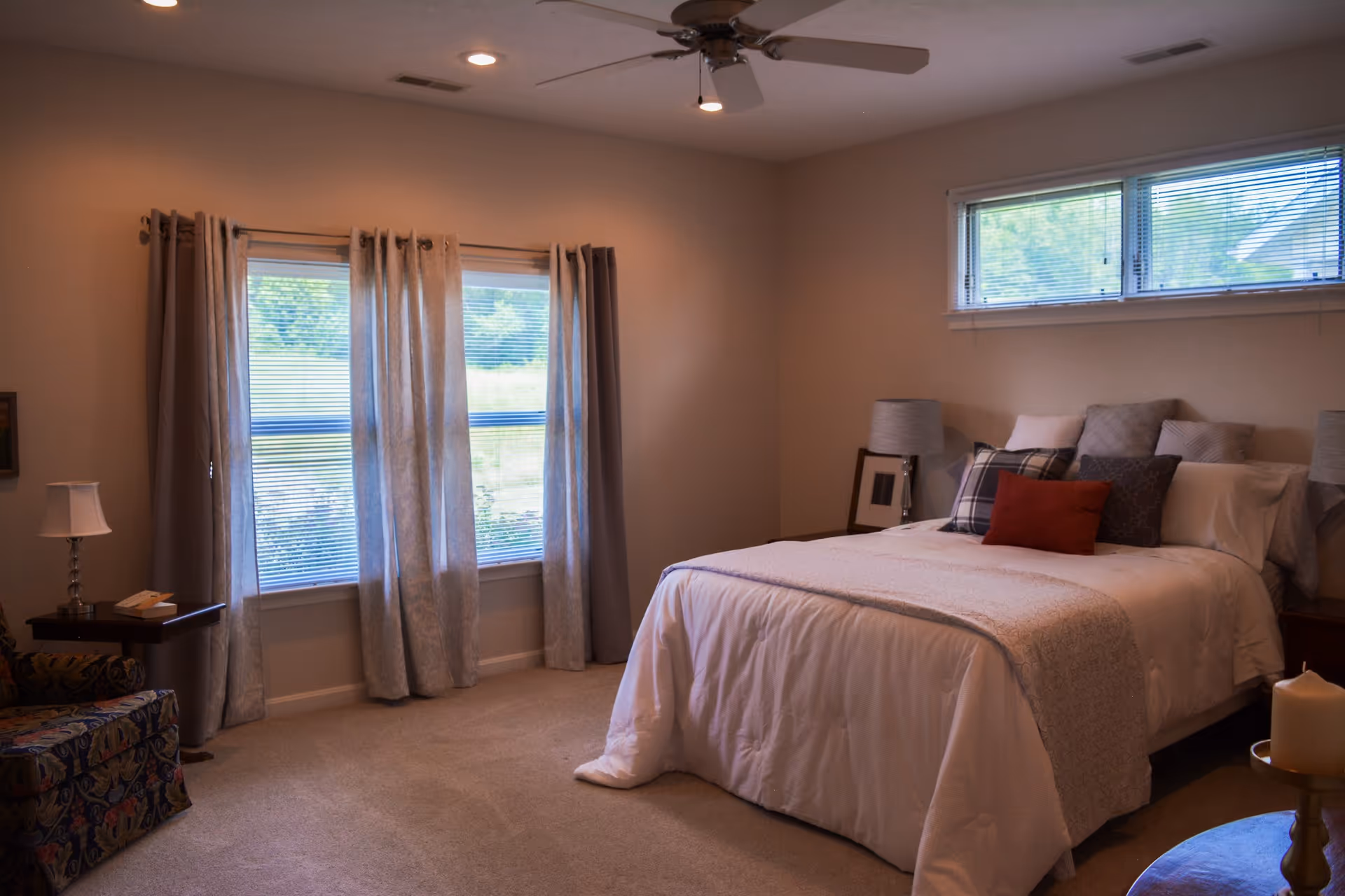 Sunlit bedroom with a made bed, decorative pillows, bedside tables and lamps, windows with curtains, and a ceiling fan.