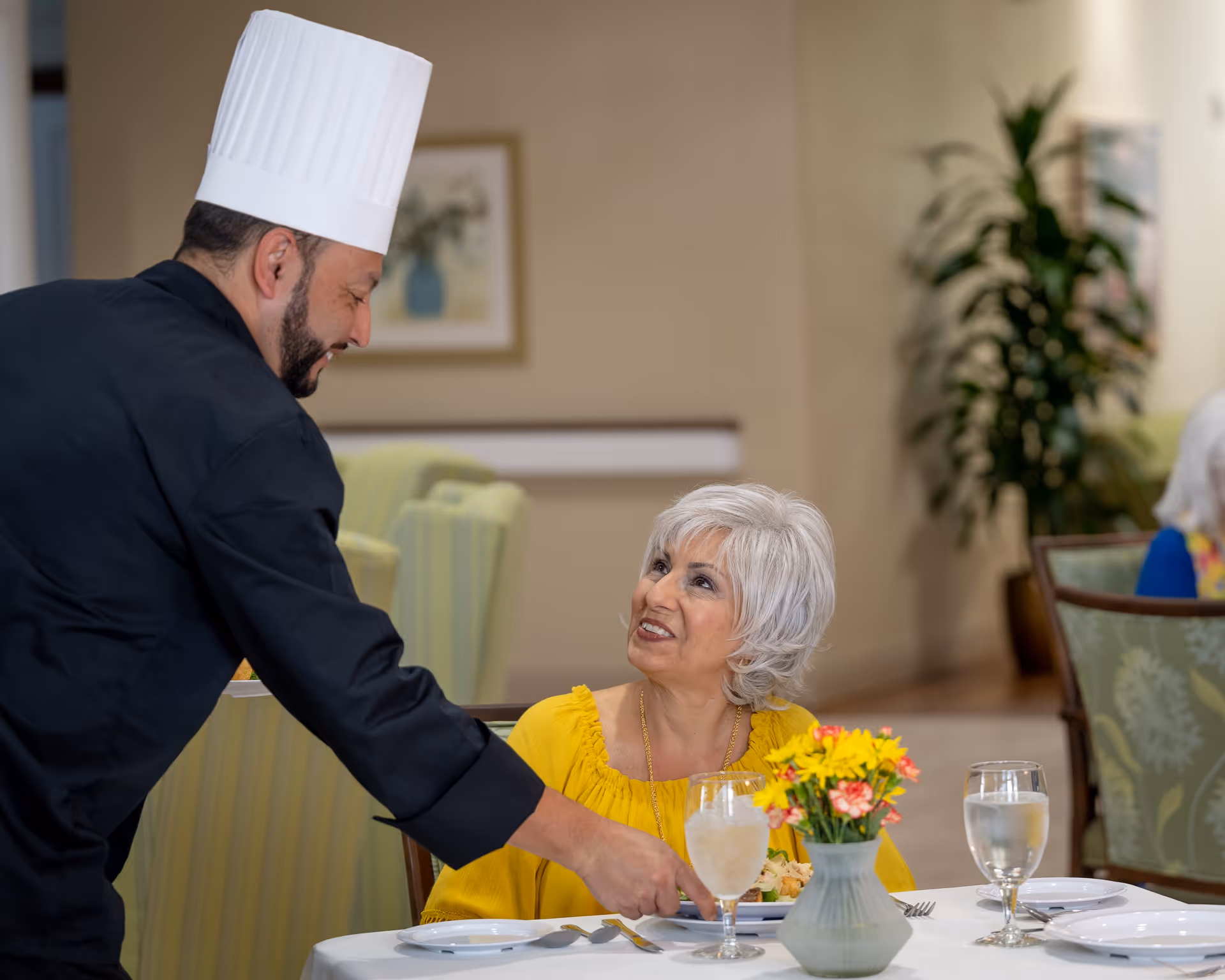 A chef in a black uniform and white chef hat serves a meal to a smiling elderly woman with white hair wearing a yellow top, seated at a dining table with glasses of water and a vase of yellow and pink flowers in a senior living facility.