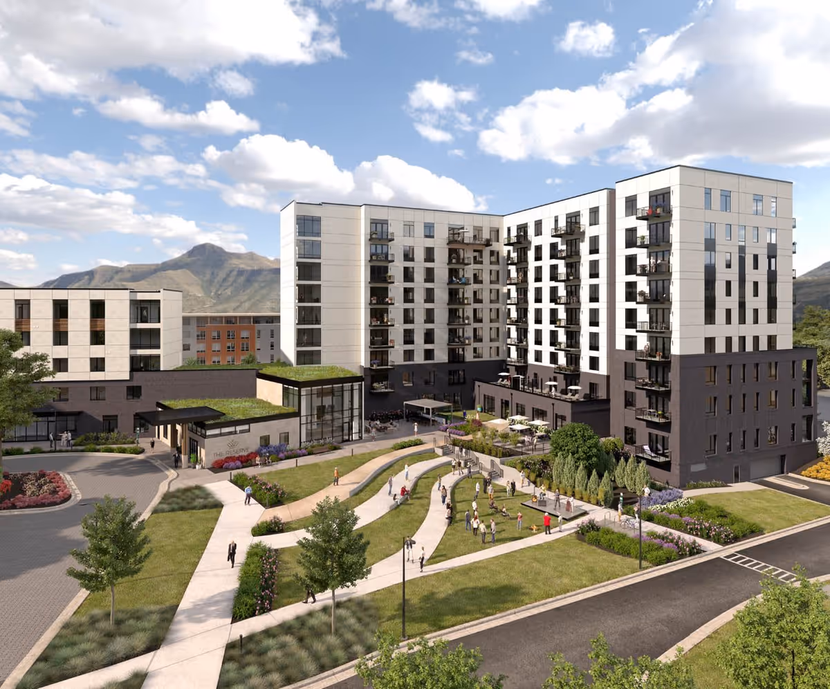 Exterior view of a modern multi-story senior living facility named The Reserve at Lone Tree, featuring landscaped gardens, walking paths, and outdoor seating areas with people enjoying the outdoor space. Mountains and a partly cloudy sky are visible in the background.