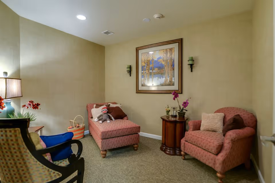 A cozy sitting area in a senior living facility with two upholstered chairs and a chaise lounge in a pink patterned fabric. There is a small round wooden table between the chaise lounge and one chair, decorated with a small plant and flowers. A framed landscape painting hangs on the beige wall above the table, flanked by two wall sconces with green candles. A basket with yarn and a sock monkey toy are placed on the chaise lounge. A lamp with a beige shade and a green base is on a side table next to another chair with a blue cushion.