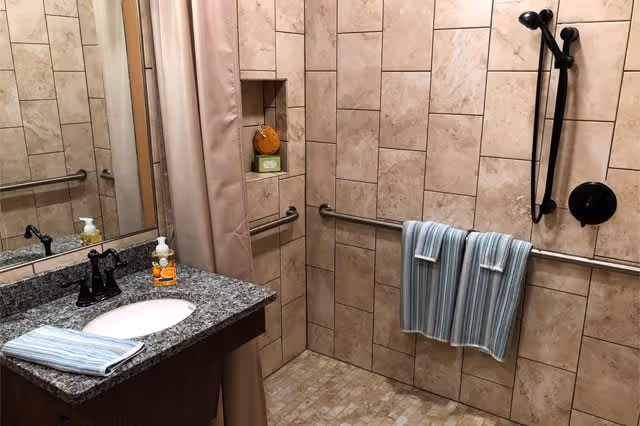 Accessible tiled bathroom with a granite sink counter, walk-in shower, grab bars, and striped towels hanging on a rail.
