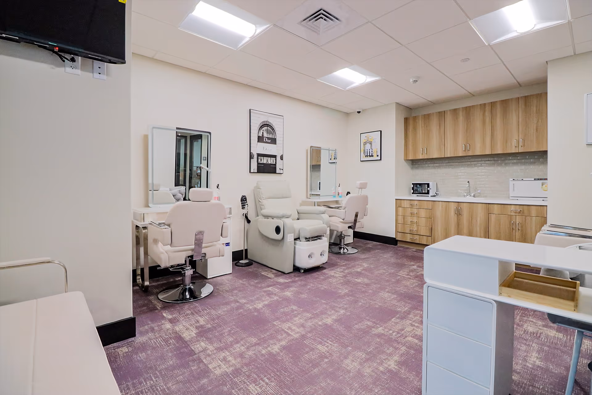 Interior room with purple carpet, two salon chairs with mirrors, a reclining chair, wooden cabinets with a microwave and sink, and a white counter in the foreground.