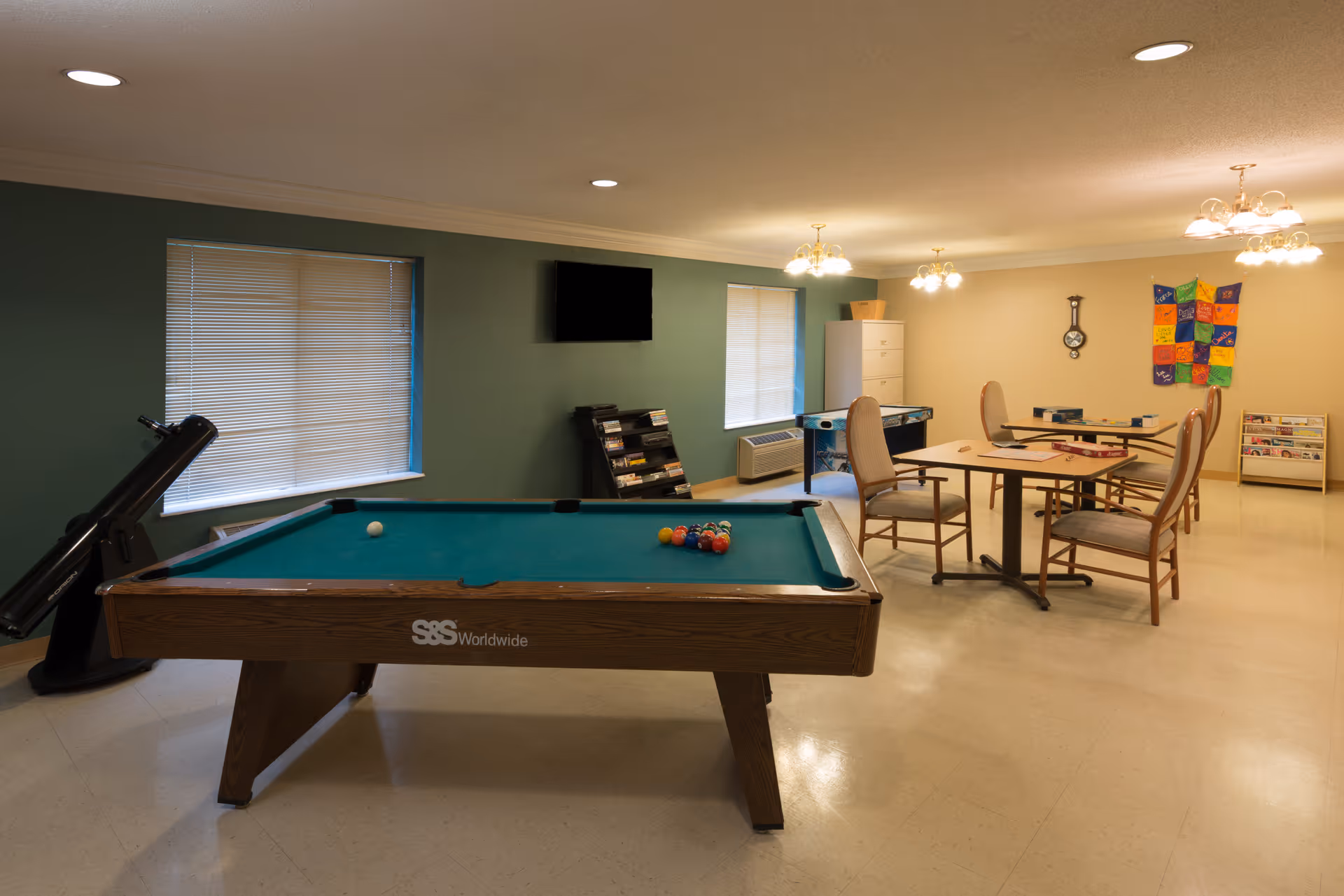 A recreational room with a pool table in the foreground, a telescope near the window, and a table with four chairs in the background. The room has green and beige walls, two windows with blinds, a wall-mounted TV, a clock, and a colorful quilt hanging on the wall. There are also shelves with magazines or books and a small air conditioning unit below one window.