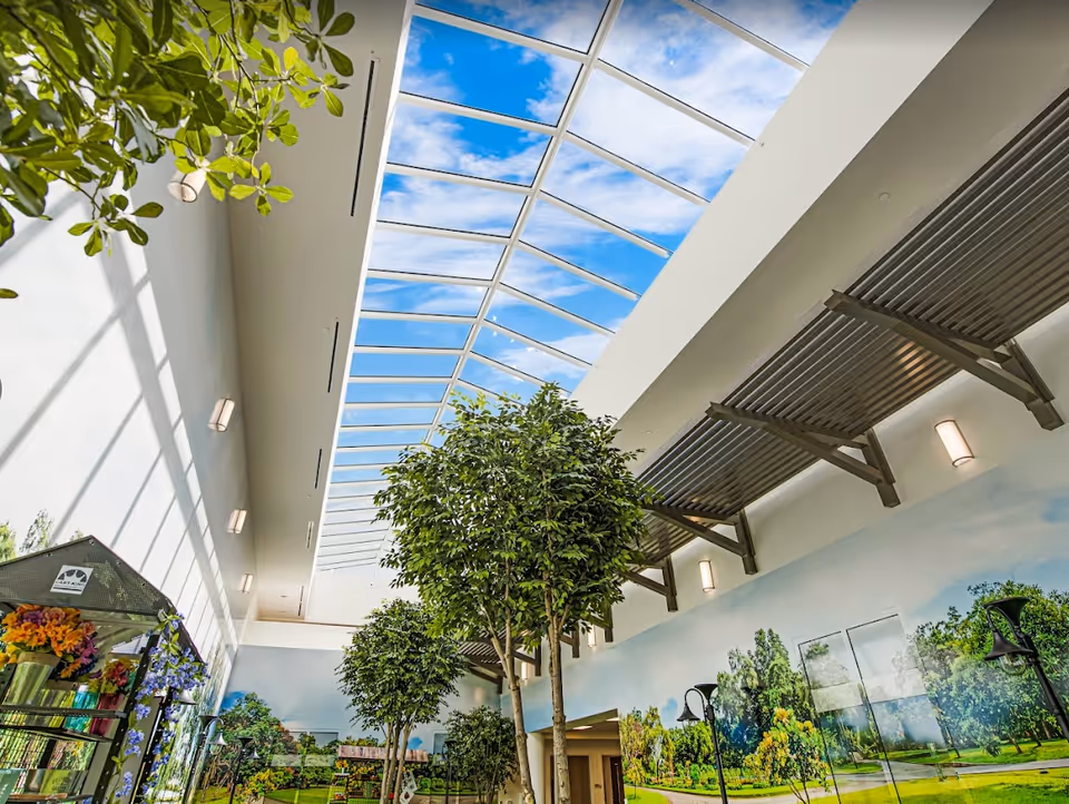 Bright indoor atrium with a long skylight, tall indoor trees and a muraled wall depicting outdoor scenery.