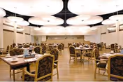A spacious dining room with multiple round and rectangular tables surrounded by cushioned chairs with patterned upholstery. The room has wooden flooring and large, circular ceiling lights providing bright illumination. The walls are light-colored with minimal decoration.