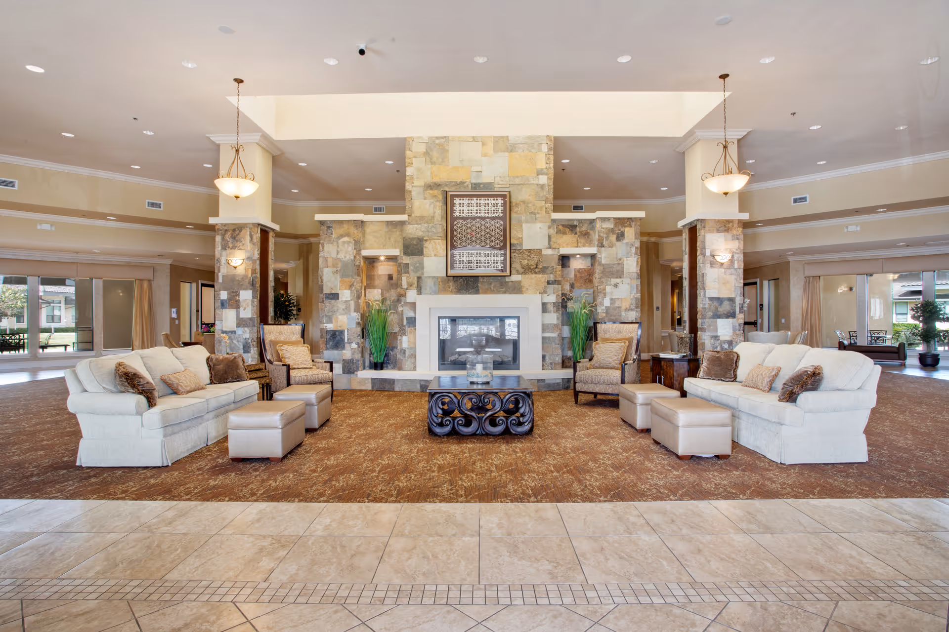 Spacious and elegant living room area in a senior living facility featuring two white sofas with brown and beige cushions, four beige armchairs, two ottomans, a decorative coffee table, and a large stone fireplace with artwork above it. The room has beige tiled flooring in the foreground and brown carpet around the seating area, with large windows and soft lighting from hanging lamps.