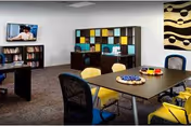 A modern common area with a table surrounded by yellow and black chairs. The table has a few decorative items on it. In the background, there is a shelving unit with colorful square compartments and a television mounted on the wall above a bookshelf. A large abstract painting is hung on the right wall.