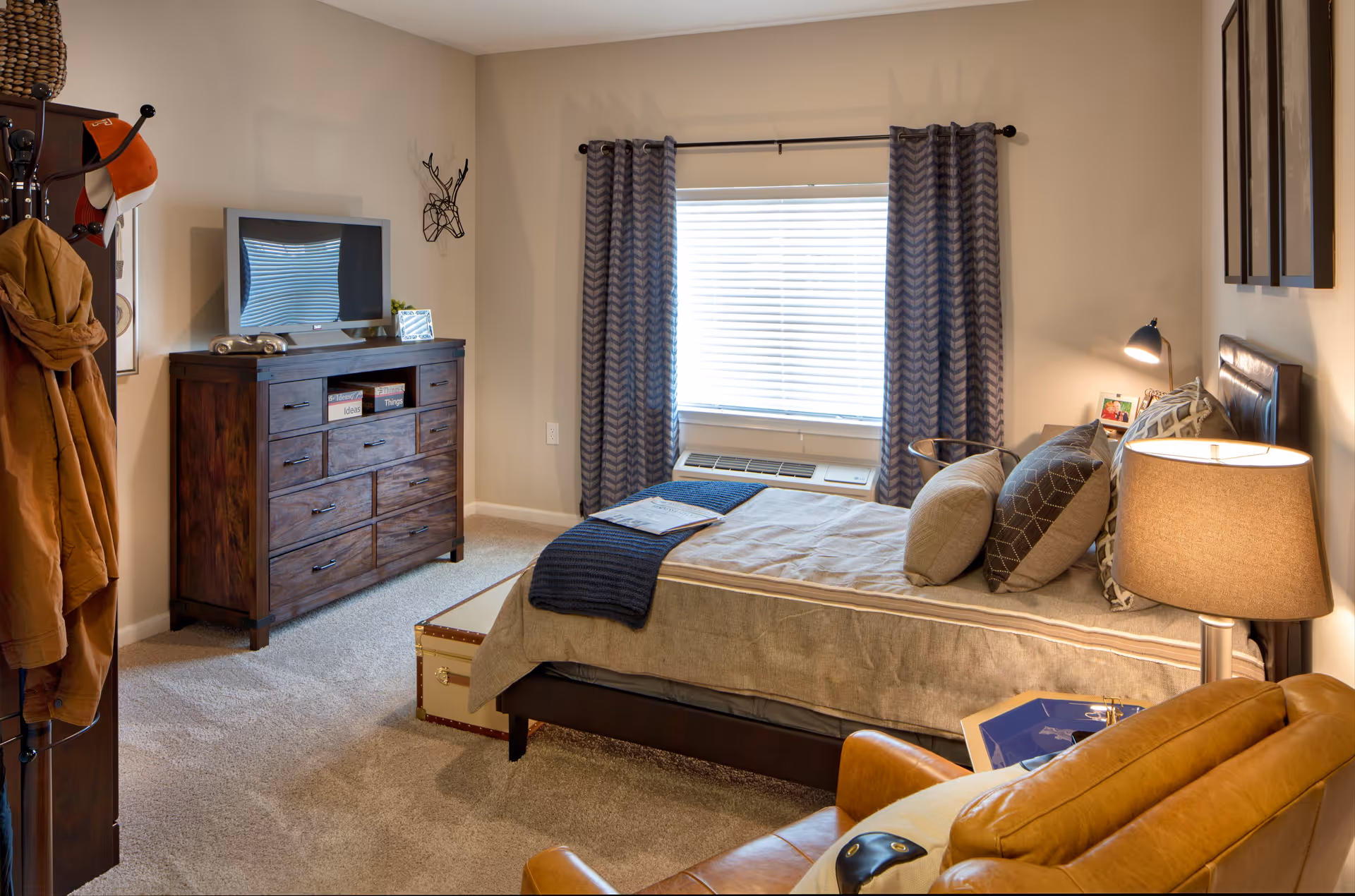 A cozy bedroom in an assisted living facility featuring a bed with multiple pillows and a beige blanket, a wooden dresser with a TV on top, a window with patterned curtains, a leather armchair, a floor lamp, and a coat rack with jackets and hats.