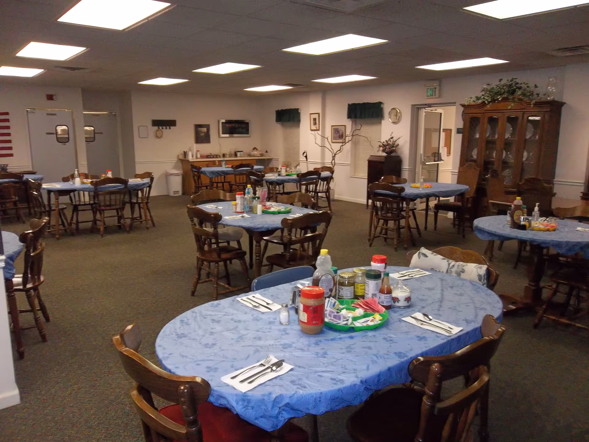 A dining room with multiple round and oval tables covered with blue tablecloths. Each table is set with utensils and condiments, including jars, bottles, and a tray with packets. Wooden chairs surround the tables. The room has carpeted floors, fluorescent ceiling lights, and a wooden cabinet with glass doors in the background.