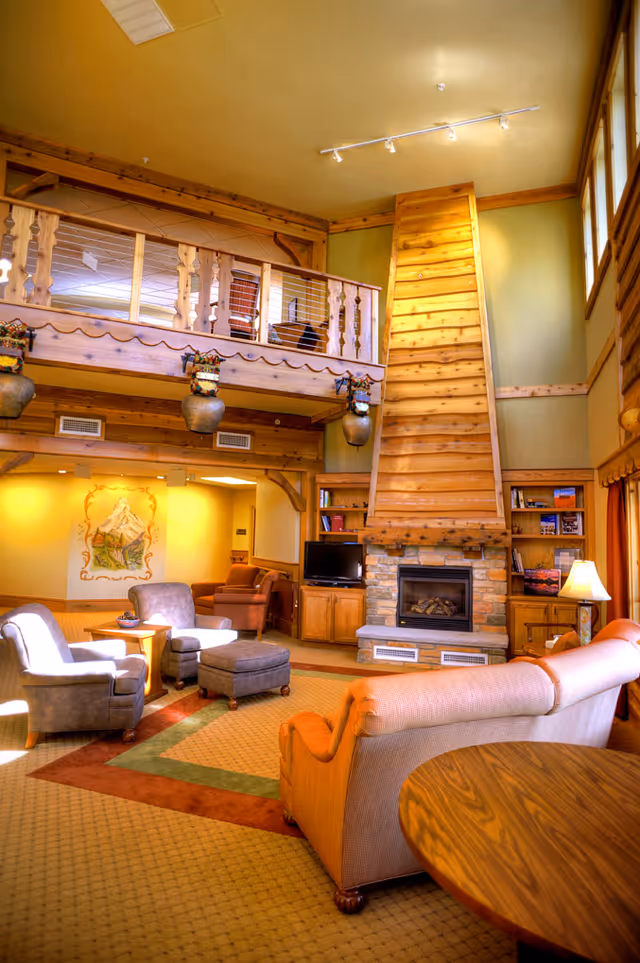 Cozy multi-level wood-paneled common room with sofas, armchairs, a stone fireplace, and an upper loft balcony.