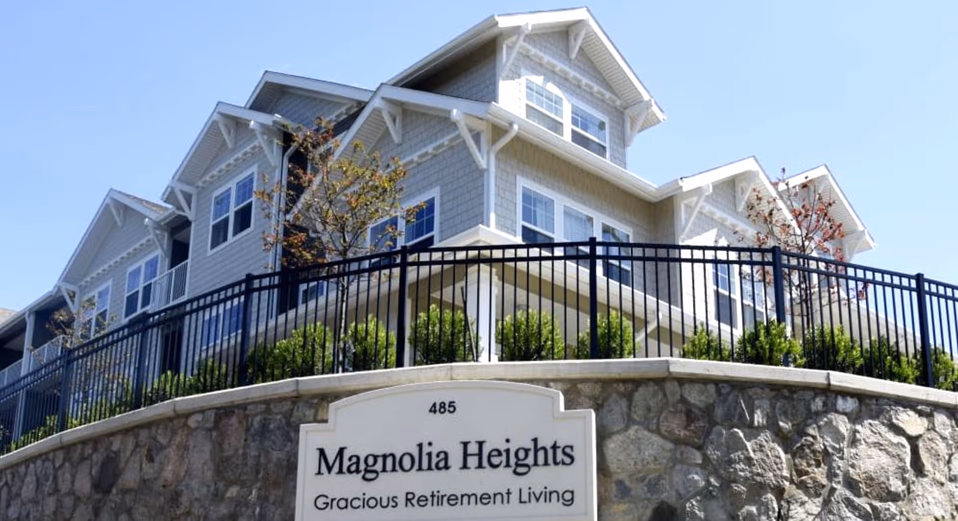 Exterior view of a multi-story residential building with gray siding and white trim, surrounded by a black metal fence and landscaping. A stone wall in front displays a sign that reads '485 Magnolia Heights Gracious Retirement Living' under a clear blue sky.
