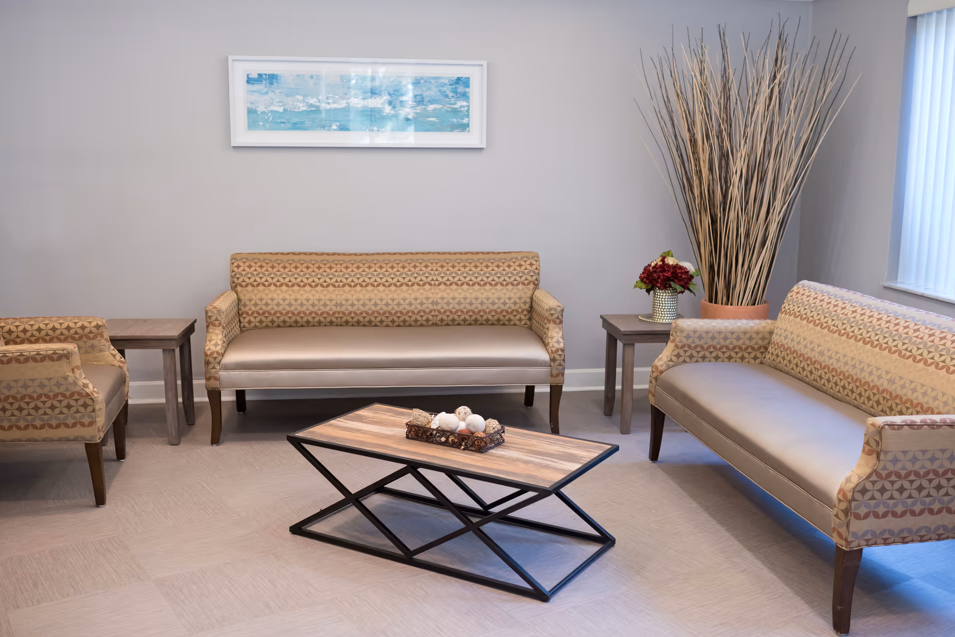 A seating area with two patterned sofas and one matching armchair arranged around a modern wooden coffee table with a black metal frame. There are two small side tables, one with a vase of flowers and the other empty. A tall decorative arrangement of dried branches is placed in the corner near a window with vertical blinds. A framed abstract blue and white painting hangs on the light gray wall.