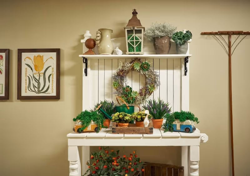 A decorative white wooden table with various potted plants, a wreath, and small ornamental items arranged on and above it against a beige wall. There are framed floral prints on the wall to the left and a wooden coat rack to the right.