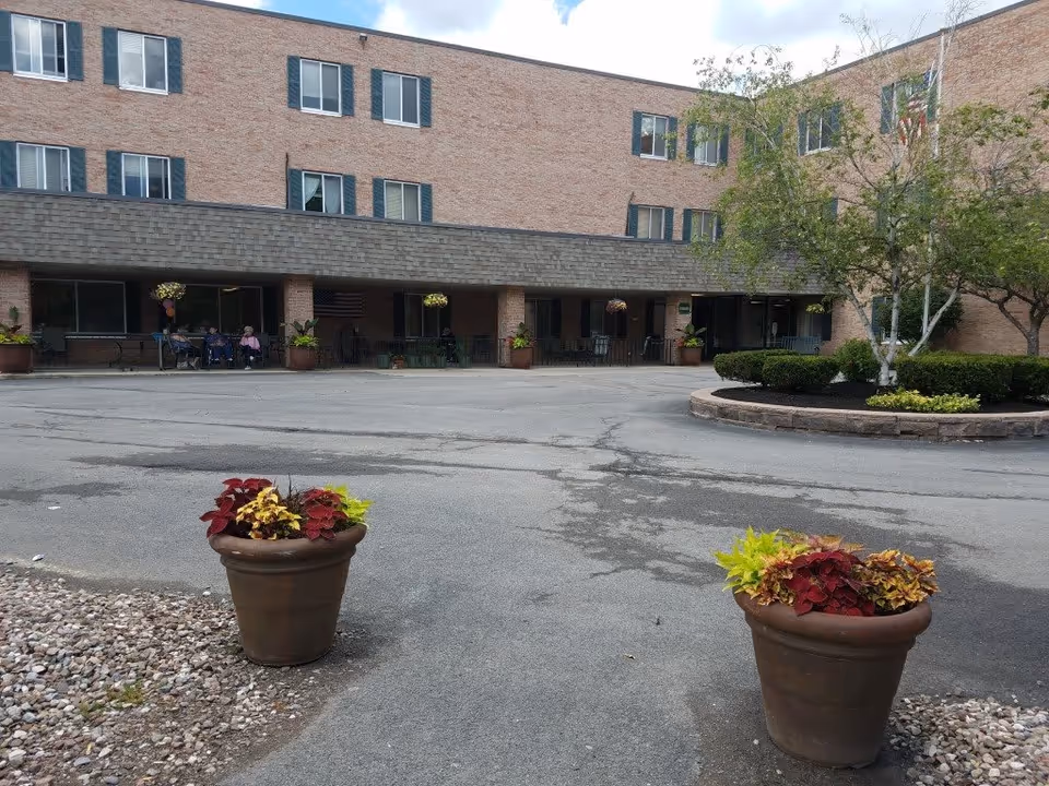 Exterior view of a three-story brick building with multiple windows and a covered patio area. There are two large flower pots with colorful plants in the foreground, a circular landscaped area with bushes and trees to the right, and a paved driveway or parking area in front of the building. A few people are sitting under the covered patio.