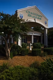 Entrance of Gaither Suites At West Park Assisted Living and Personal Care Home with a white archway displaying the facility name, surrounded by green bushes and trees under a clear blue sky.