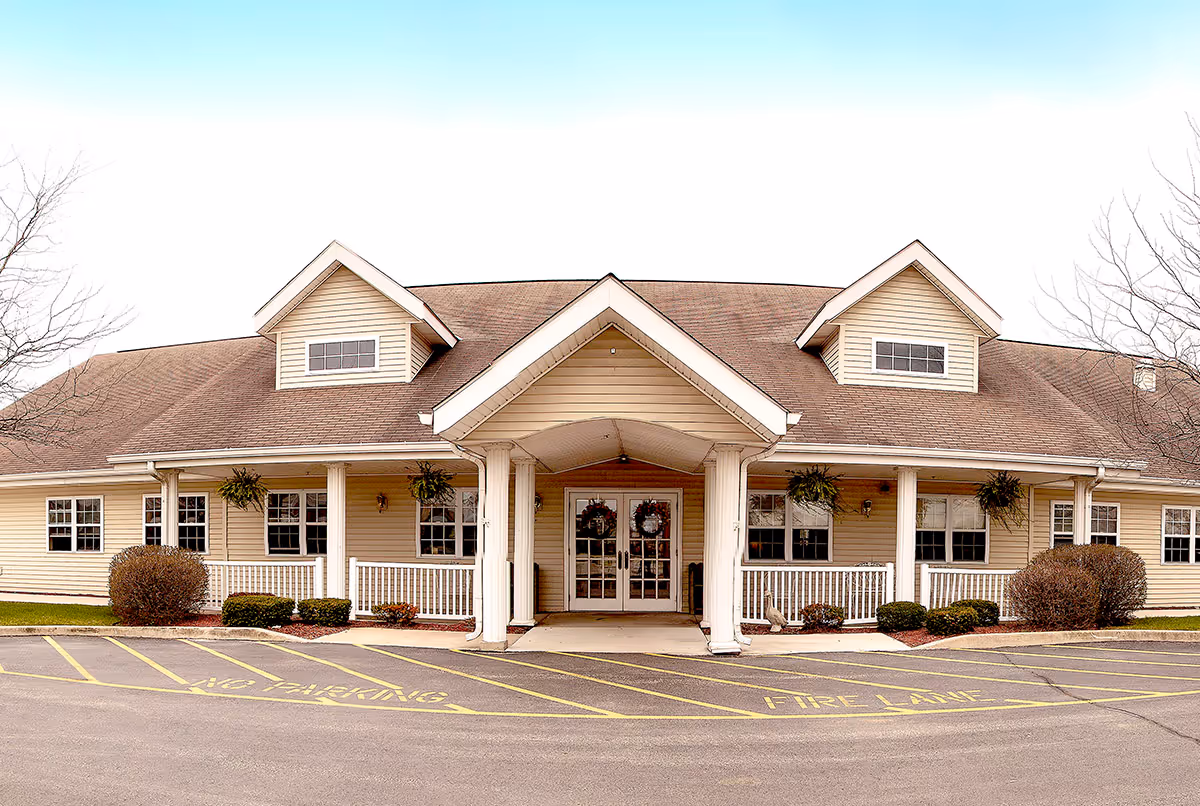 Front exterior view of a single-story building with beige siding and a brown roof. The entrance features a covered porch with white columns and double glass doors decorated with wreaths. There are several windows along the front, hanging plants on the porch, and neatly trimmed bushes. The parking area in front has yellow painted markings indicating no parking and fire lane.