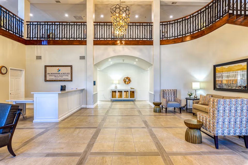 Spacious senior living facility lobby with a reception desk, seating area, chandelier, and upstairs balcony railing.