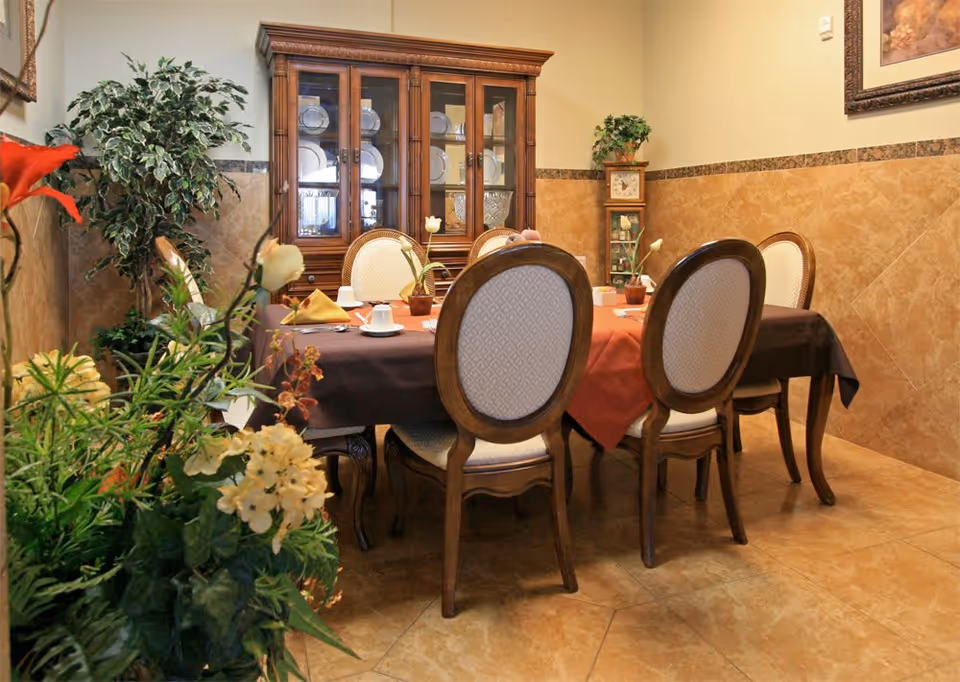 A dining room with a wooden dining table covered with a brown and orange tablecloth, surrounded by six upholstered chairs with wooden frames. The table is set with cups, napkins, and small flower vases. In the background, there is a wooden china cabinet displaying plates and glassware. The room has beige tiled walls and floor, with decorative plants on the left side and a small wooden shelf with a plant on top in the corner.