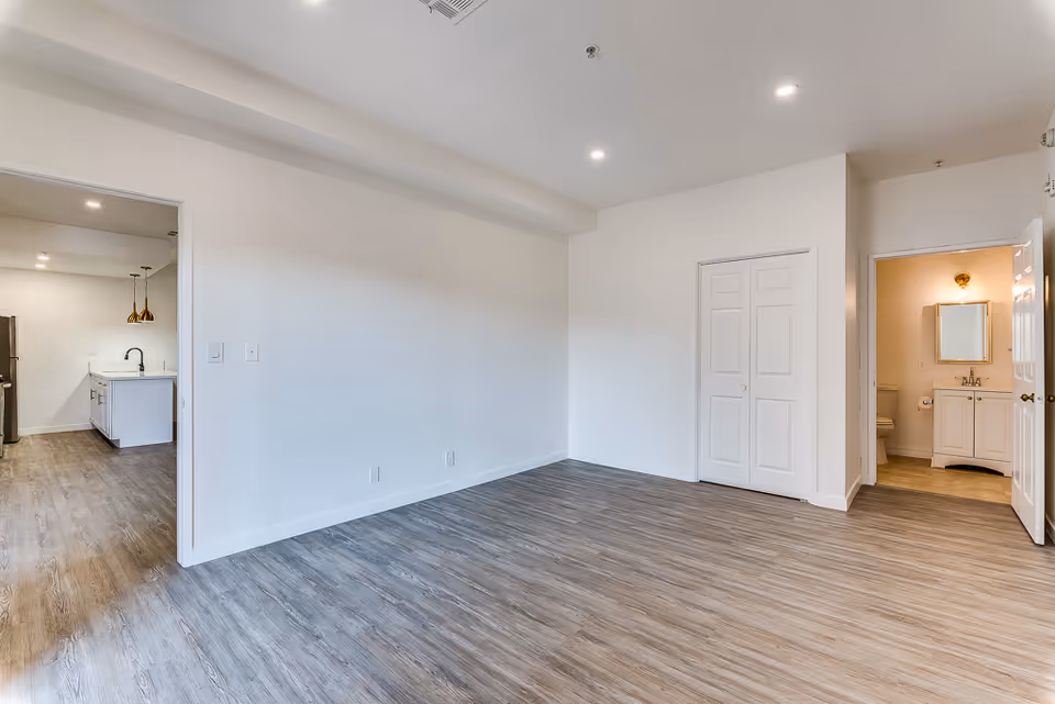 Empty room with wood-style flooring and white walls, featuring a closed closet with double doors and an open doorway leading to a bathroom with a sink, mirror, and toilet. Another open doorway leads to a kitchen area with white cabinets, a black faucet, and pendant lights.