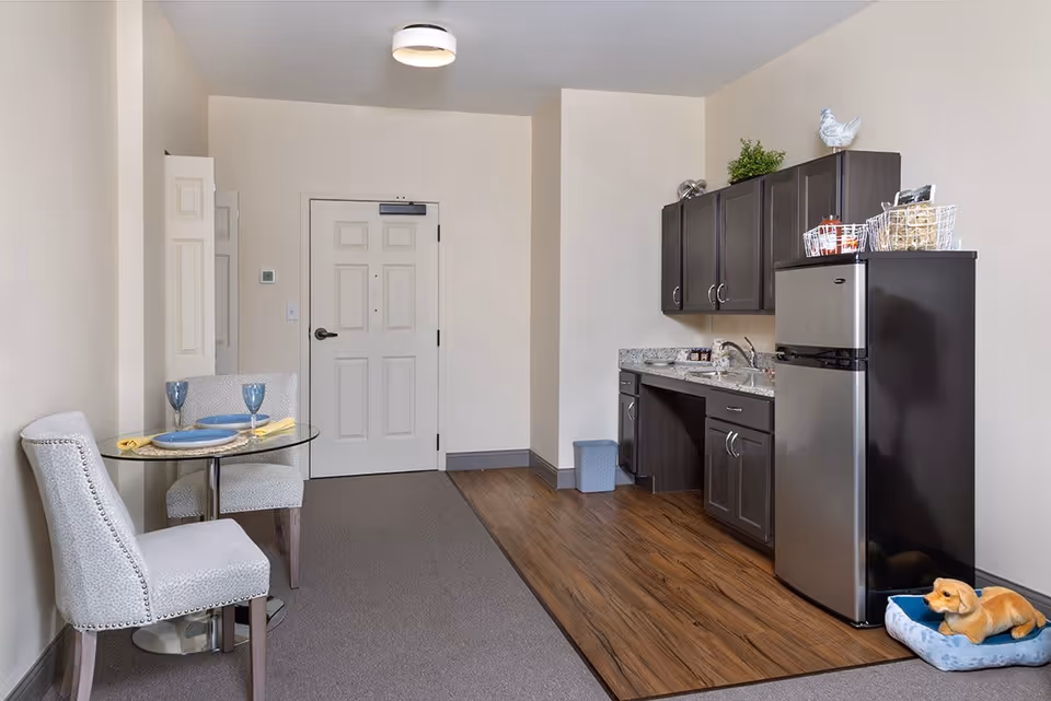 A small kitchenette area in a senior living facility with dark cabinets, a stainless steel mini refrigerator, and a granite countertop with a sink. To the left, there is a small round glass dining table set with two chairs, two blue plates, two blue glasses, and yellow napkins. The floor is a combination of wood and carpet, and a dog bed with a small dog figurine is placed near the refrigerator. The walls are light beige, and there is a white door in the background.