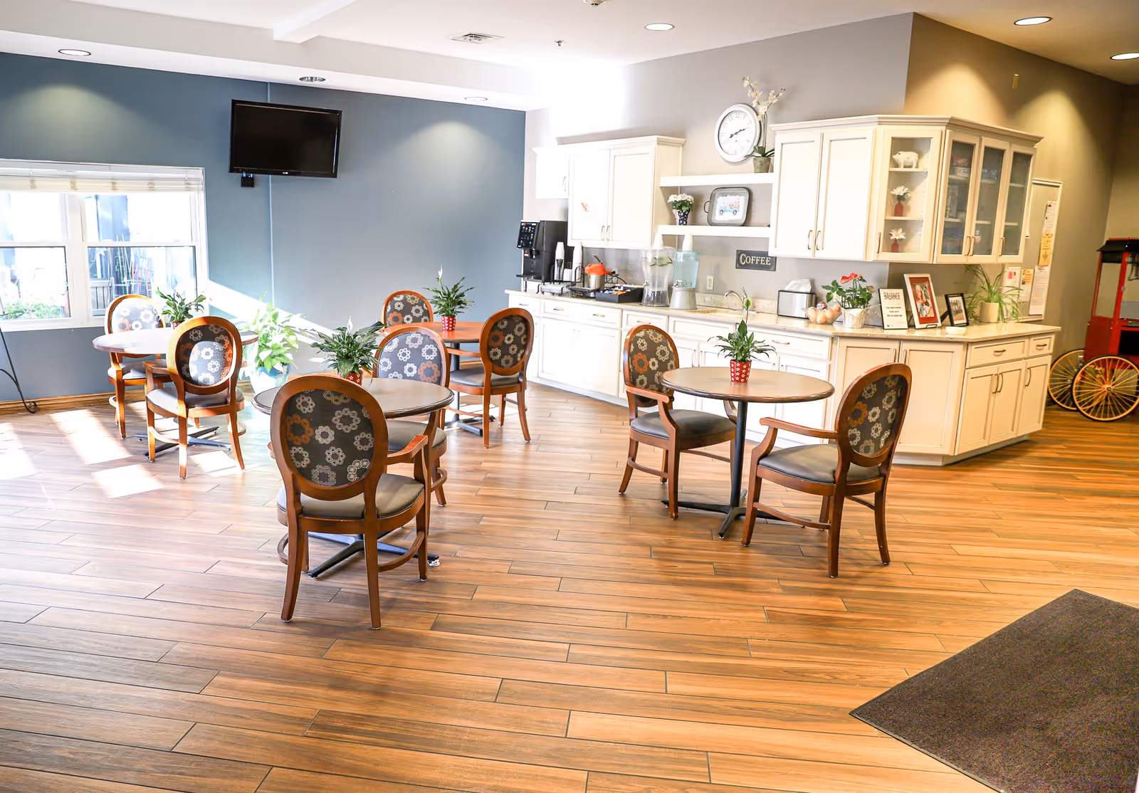 A bright and clean dining area in a senior living facility with several round wooden tables and cushioned chairs with floral upholstery. The room has wood-look flooring, a blue accent wall with a mounted flat-screen TV, and a white kitchenette area with cabinets, a coffee station, and decorative plants and pictures.