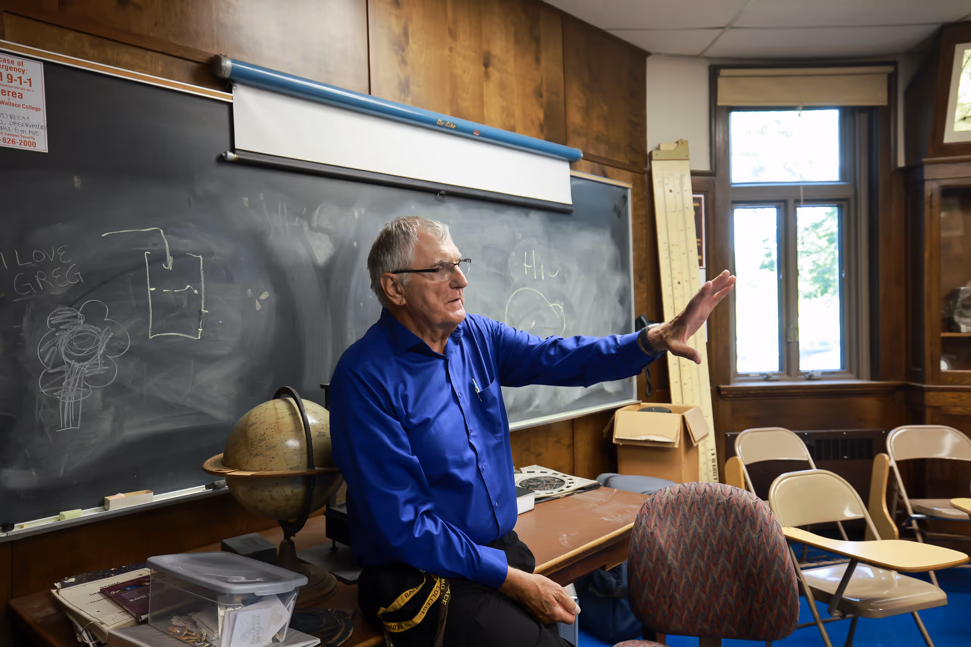 An elderly man wearing a blue shirt is sitting on a desk in a classroom with wooden paneling, a blackboard with chalk drawings, a globe, and several empty chairs with attached desks. There is a window letting in natural light and some classroom supplies on the desk.