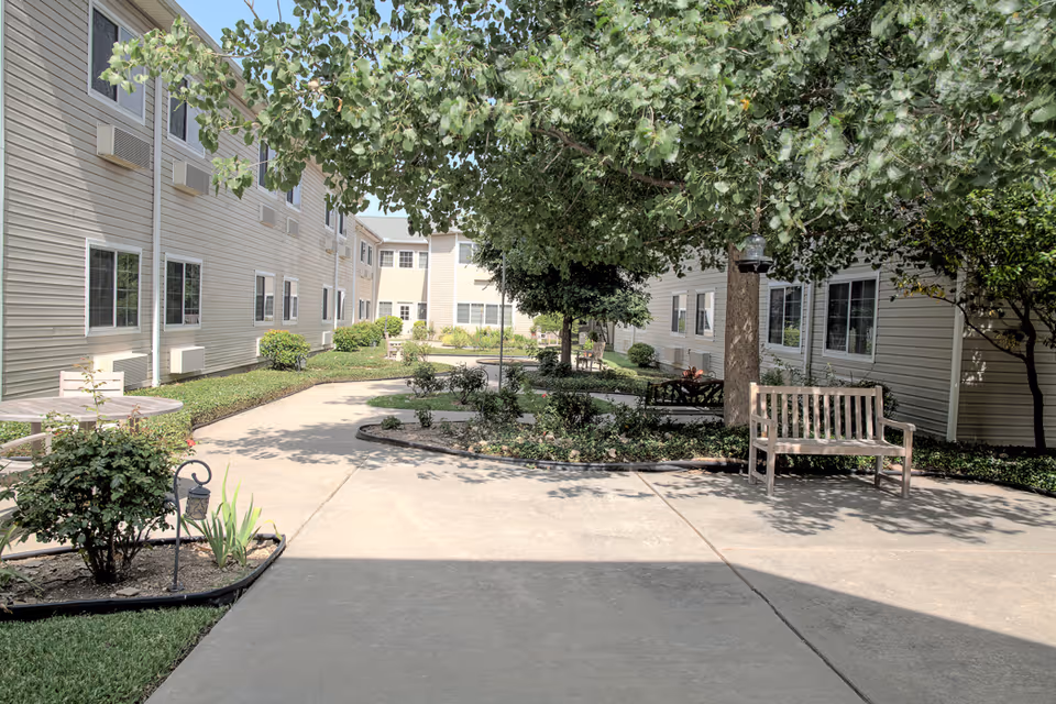 Outdoor courtyard area at Garden Estates of Temple featuring paved walkways, benches, trees, and shrubs surrounded by beige multi-story building walls with windows and air conditioning units.