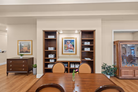 Wood dining table and chairs in a common room facing built-in shelving, framed artwork, and cabinets.