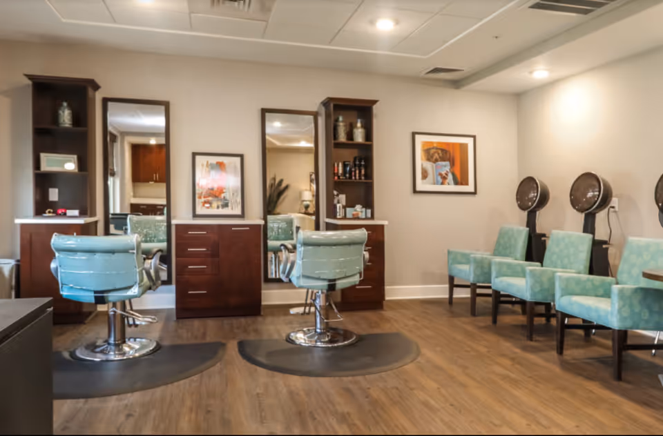 Bright salon room with two styling stations, teal salon chairs, mirrors, and several waiting chairs with hooded hair dryers.