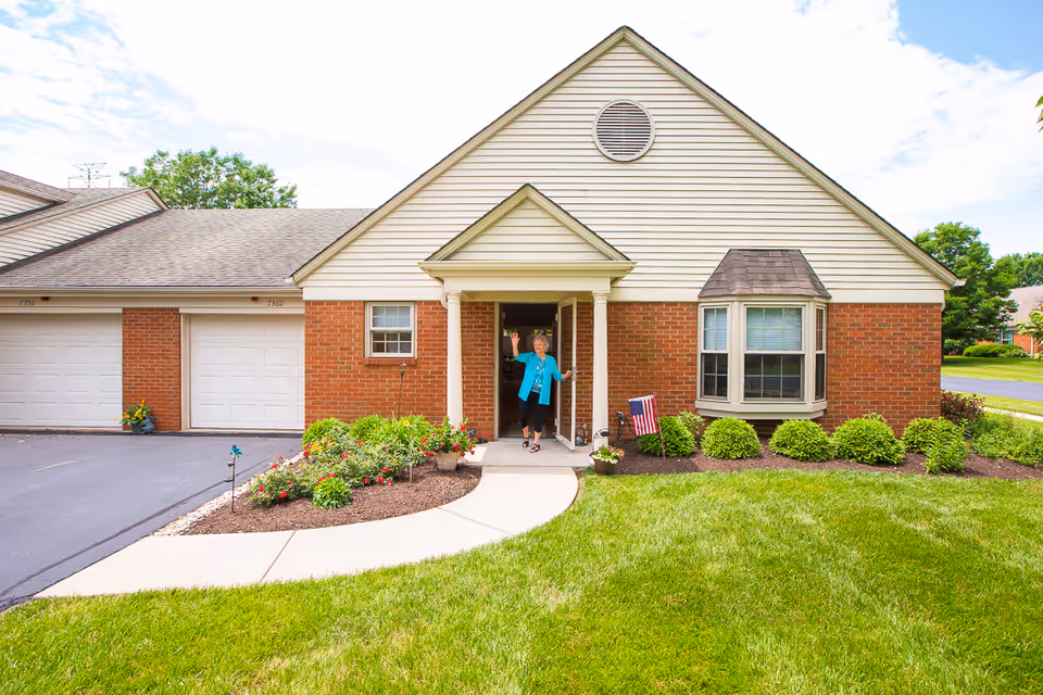 A single-story brick and siding house with a small covered porch. A woman in a blue jacket is standing in the doorway waving. The house has a well-maintained lawn, flower beds, and an American flag near the porch. There are two white garage doors on the left side of the house.