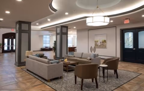 A spacious and well-lit living room area in The Remington of Yardley featuring two gray sofas, two brown armchairs, a wooden coffee table, and a patterned rug. The room has decorative columns, recessed ceiling lights, a modern chandelier, and framed artwork on the wall. There are large windows and double doors visible in the background.