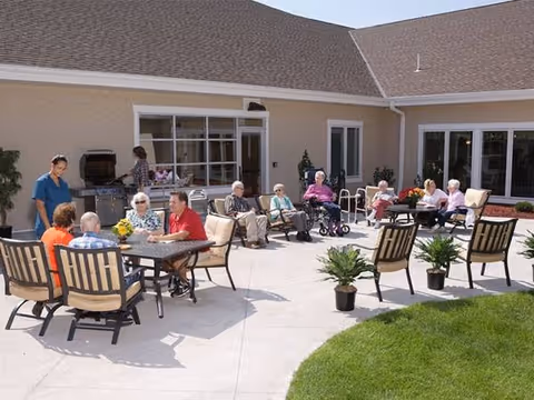 A group of elderly people sitting and socializing outdoors on a sunny patio area with tables, chairs, and potted plants. A caregiver stands nearby. The patio is adjacent to a beige building with large windows and doors.