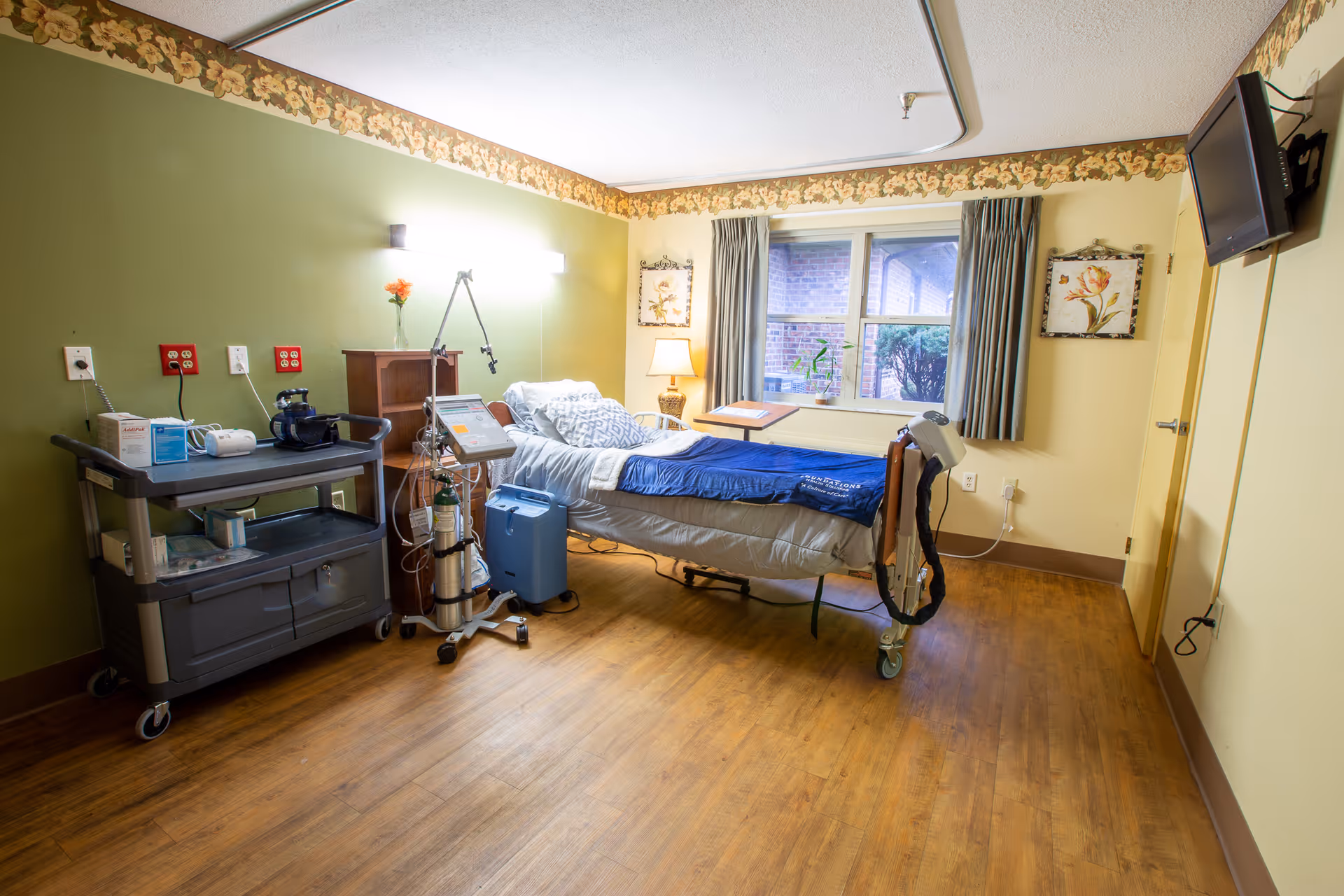 Hospital-style bedroom with an adjustable bed, medical cart and oxygen equipment beside a window.
