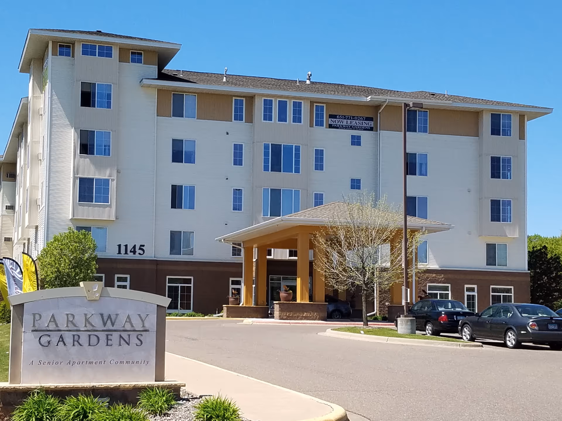 Front exterior of the Parkway Gardens senior apartment building showing the entrance canopy, driveway, parked cars, and a stone sign in the foreground.