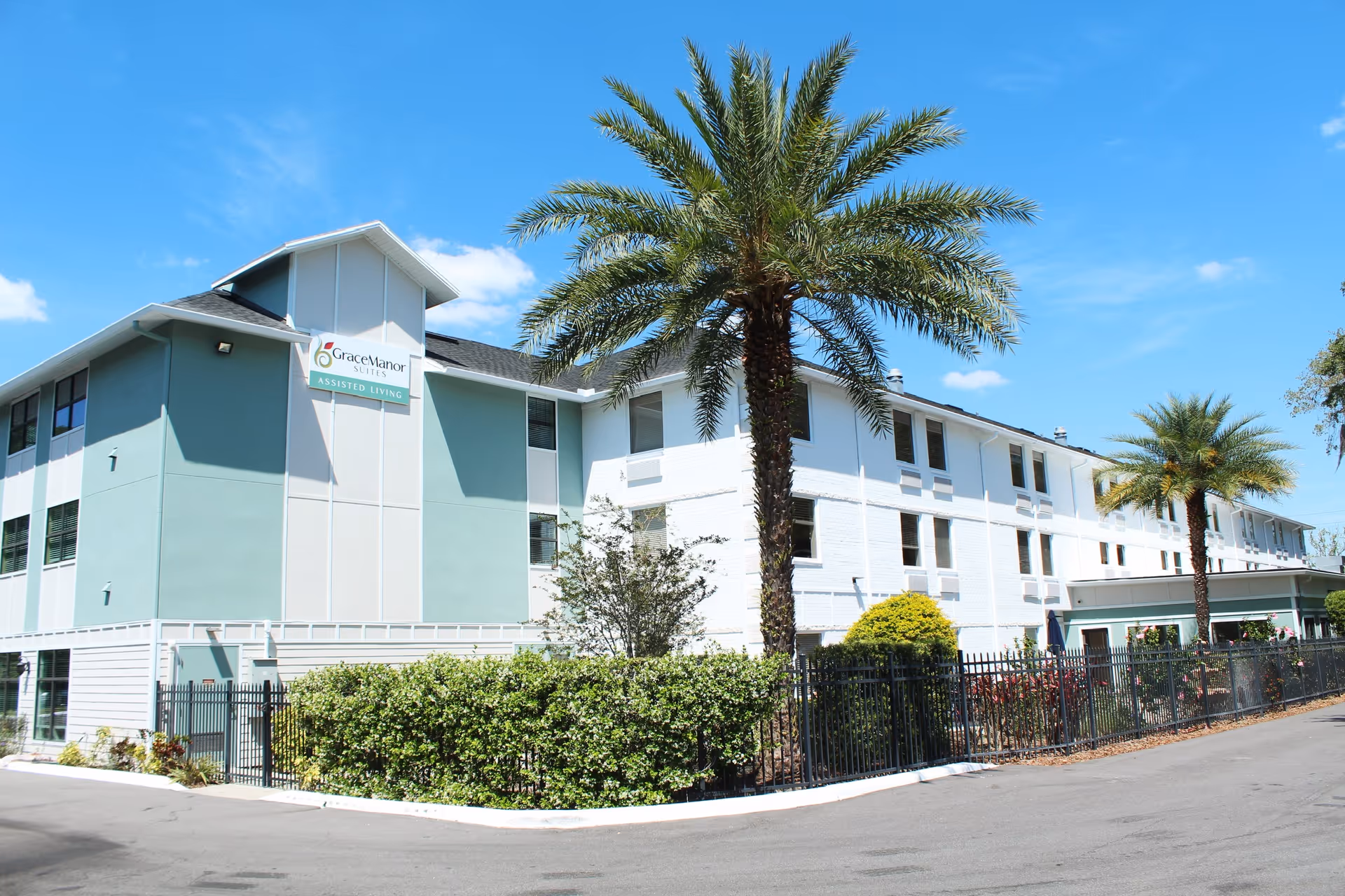 Front exterior of the Grace Manor Suites assisted living building with palm trees, landscaping, and a fenced entrance under a blue sky.