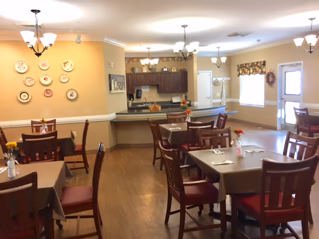 Communal dining room with several tables and chairs, floral centerpieces, and a serving counter with cabinetry in the background.