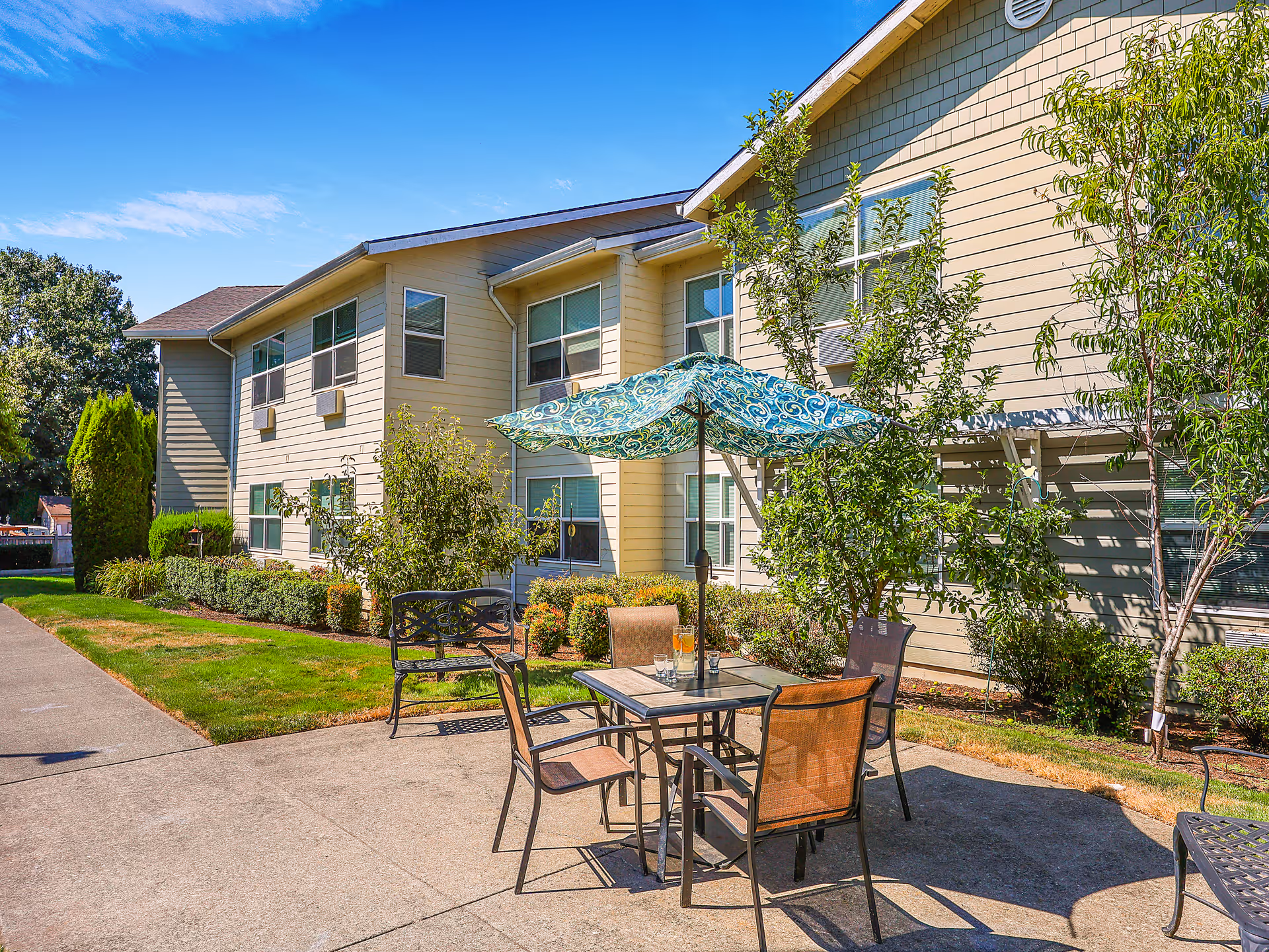 Outdoor patio area at Prestige Senior Living Auburn Meadows featuring a table with four chairs and a large blue patterned umbrella. The patio is adjacent to a two-story beige building with multiple windows, surrounded by green shrubs, trees, and a well-maintained lawn under a clear blue sky.