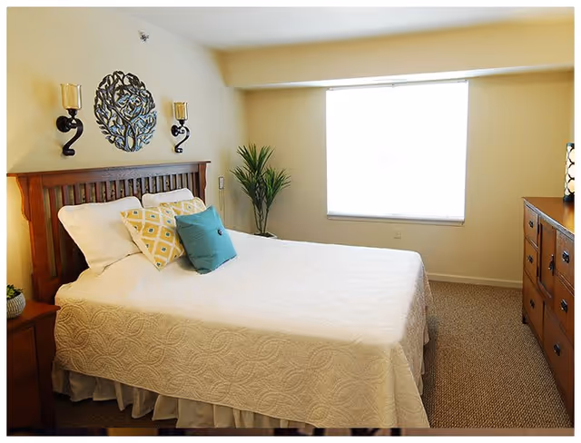 A bright bedroom with a neatly made bed featuring a white quilt and decorative pillows in yellow and teal. The room includes a wooden headboard, two wall-mounted candle holders, a metal wall decoration above the bed, a potted plant near a large window with closed blinds, and a wooden dresser with a lamp on top.