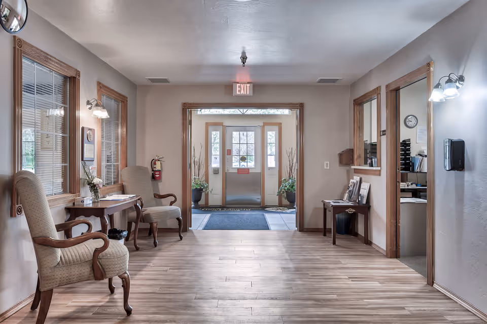 A bright and clean facility entrance area with wooden flooring, two upholstered armchairs and a small table with flowers on the left, a fire extinguisher mounted on the wall, and a reception window on the right. The exit door is visible straight ahead with glass panels and potted plants on either side. Wall-mounted lights illuminate the space.