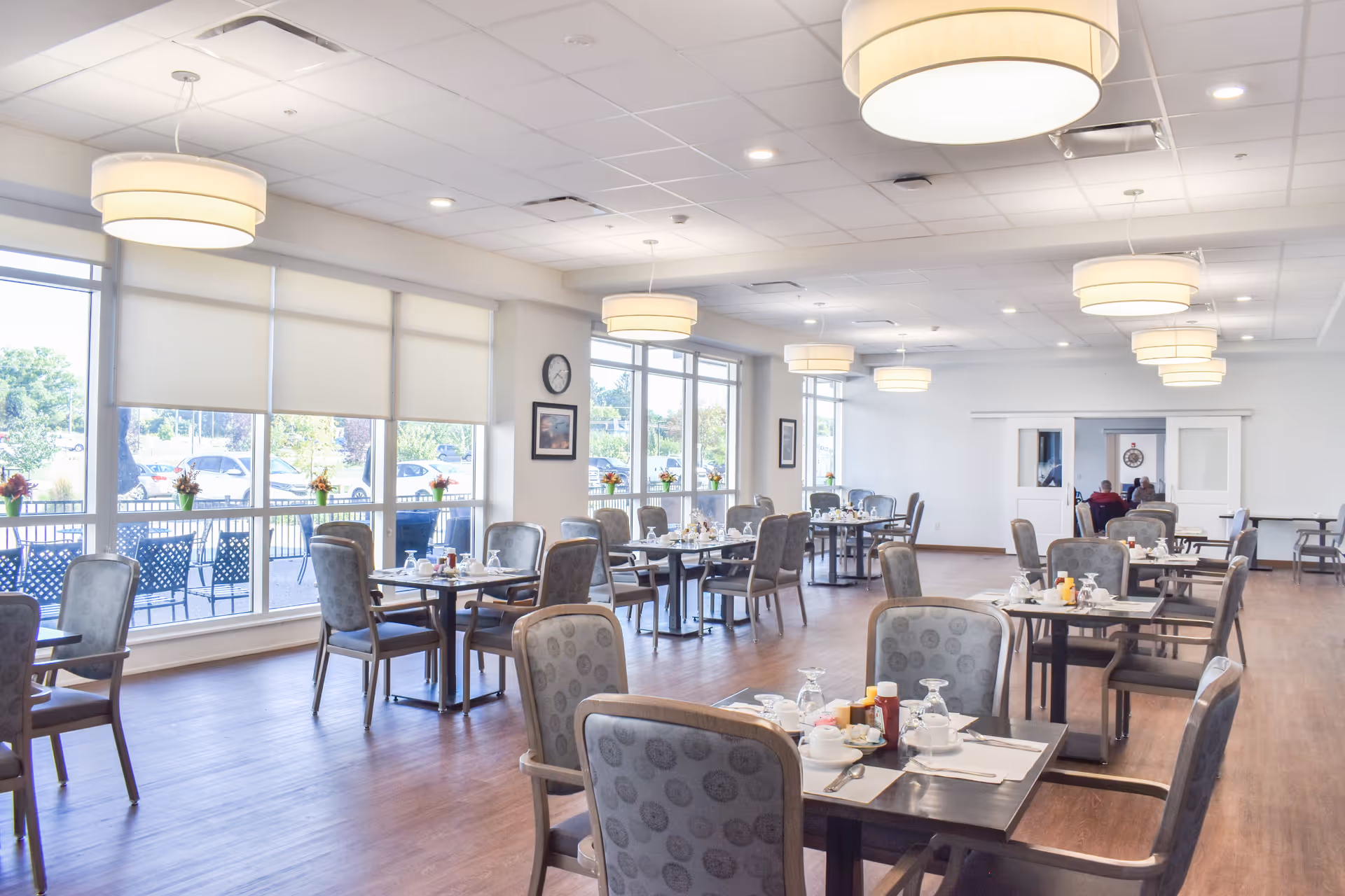 A bright and spacious dining room with multiple tables set for meals, each with chairs, placemats, utensils, and condiments. Large windows along one wall let in natural light and offer a view of the outdoor patio area with additional seating. Modern ceiling lights hang from the white ceiling, and the floor is a warm wood tone.