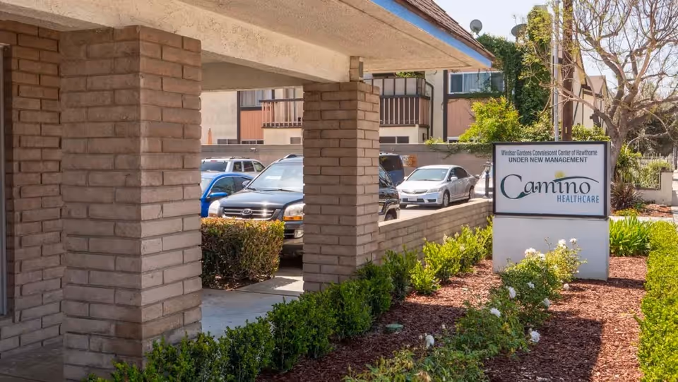 Exterior view of Camino Healthcare facility showing a brick pillar structure, parked cars, and a landscaped area with bushes and flowers. A sign reads 'Windsor Gardens Convalescent Center of Hawthorne UNDER NEW MANAGEMENT Camino Healthcare.'