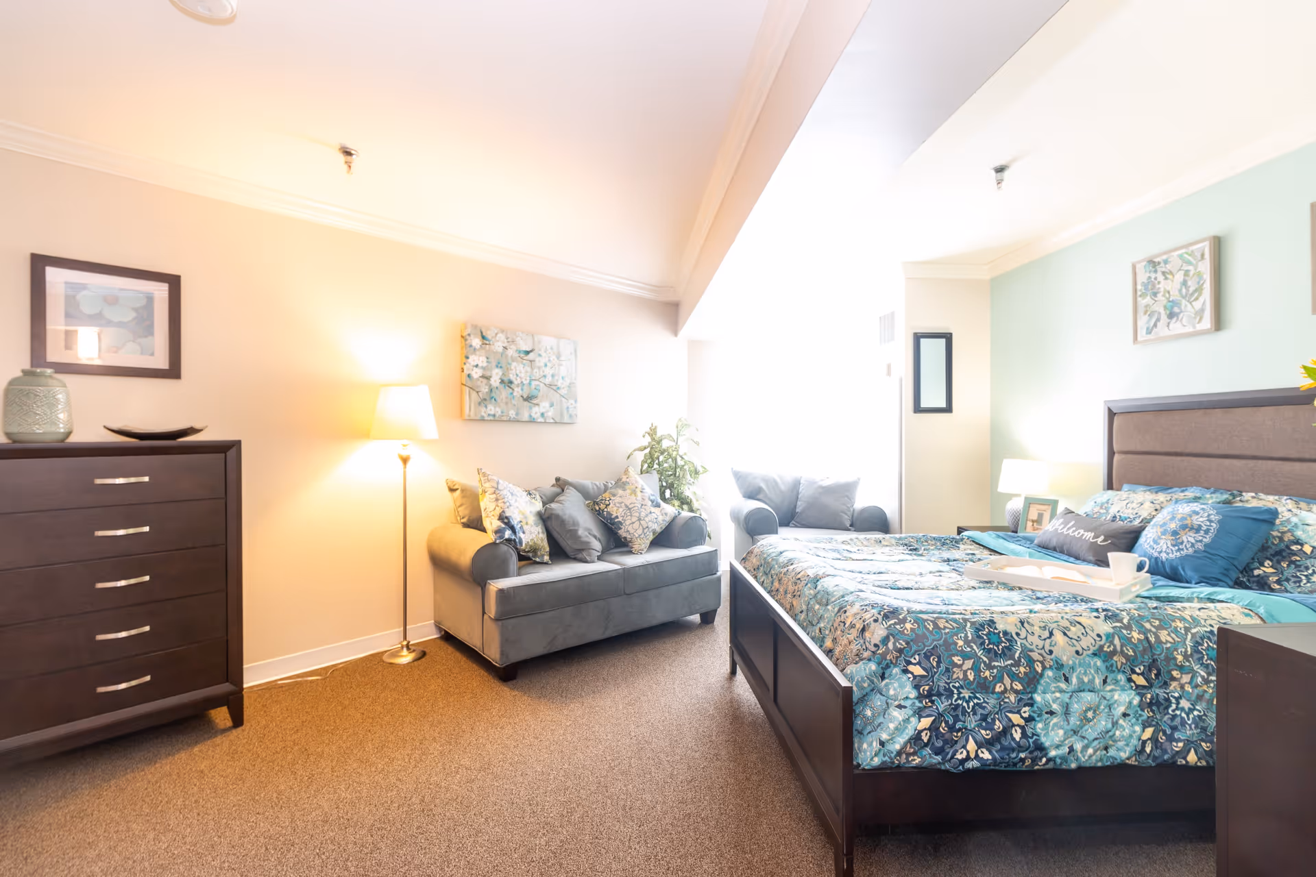 A bright and cozy bedroom in a senior living facility featuring a large bed with blue and teal patterned bedding, a dark wooden dresser, a gray loveseat with decorative pillows, a floor lamp, and framed artwork on the walls.