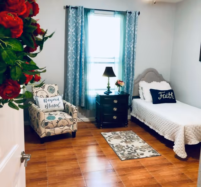 A cozy bedroom with a single bed covered in a white textured blanket and a navy blue pillow with the word 'Faith'. Next to the bed is a black nightstand with three drawers, a lamp, and a small flower vase. A patterned armchair with a pillow that says 'Beyond Blessed' is positioned near a window with blue and white curtains. The floor is wooden with a small floral rug in front of the bed. A bouquet of red roses is partially visible on the left side of the image.