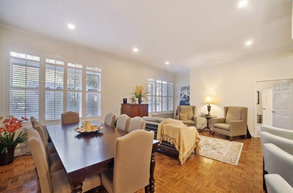 Open-plan dining and living area with a large dark wood dining table surrounded by upholstered chairs, parquet floors, and a seating area with armchairs and a lamp.