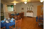 A dining room with wooden floors and several round tables covered with blue tablecloths. Each table is set with plates, glasses, and napkins. Wooden chairs surround the tables. The room has light-colored walls with a white cabinet and framed artwork on the far wall. Ceiling lights and hanging green plants provide illumination and decoration.
