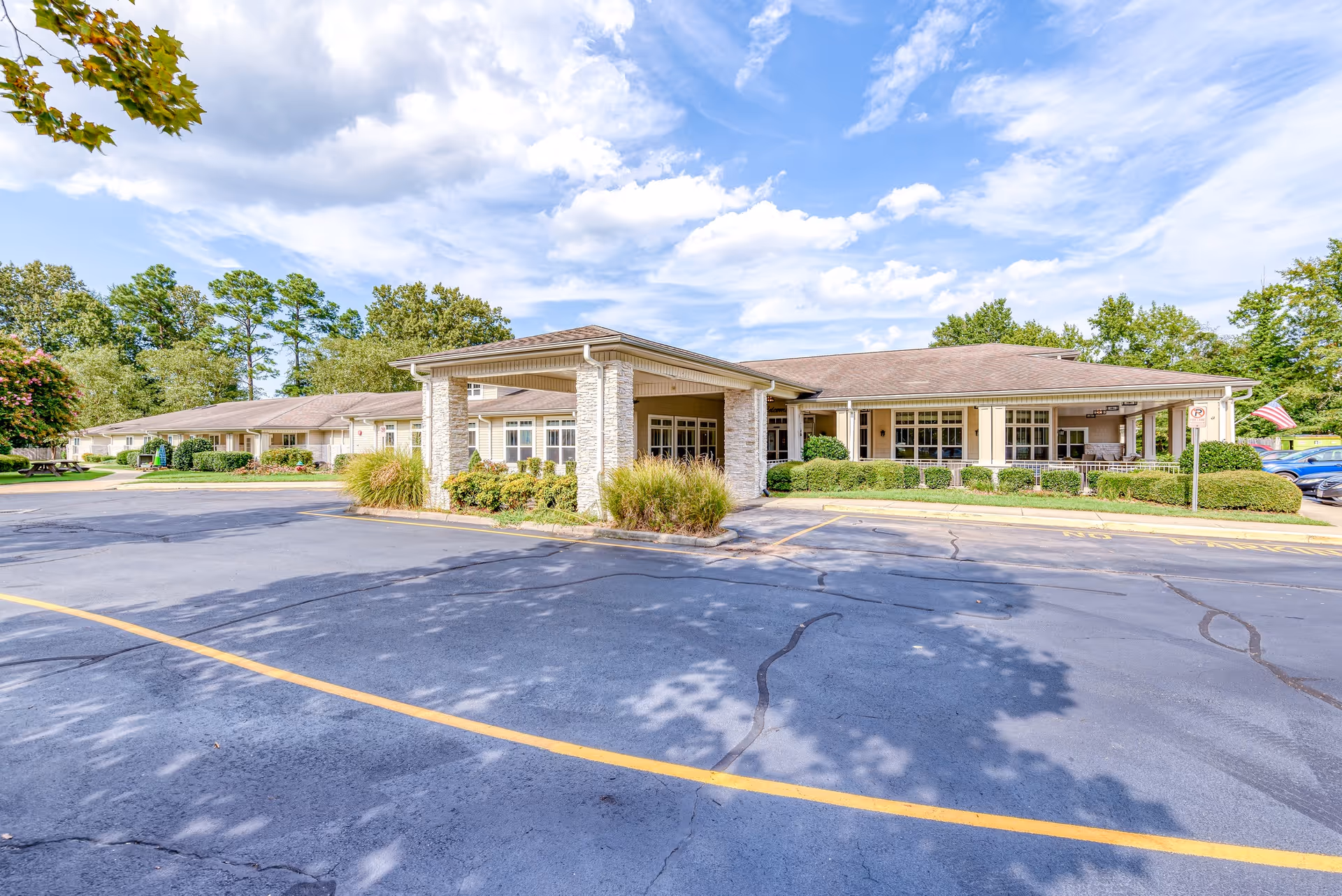 Front exterior of the Chesapeake Place Senior Living building with a covered entrance, parking lot and landscaped grounds under a partly cloudy sky.