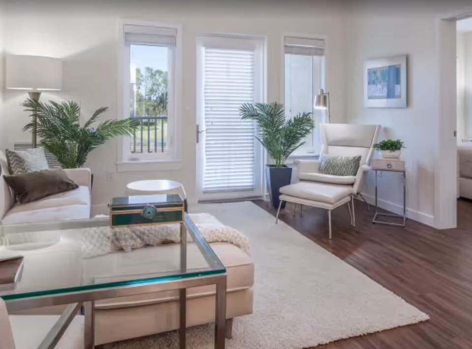 A bright and modern living room with a beige sofa adorned with pillows, a glass coffee table, a white armchair with a matching ottoman, two large potted plants, a floor lamp, and a side table with a small plant. The room has large windows and a glass door with blinds, allowing natural light to fill the space. The floor is wooden with a light-colored area rug.