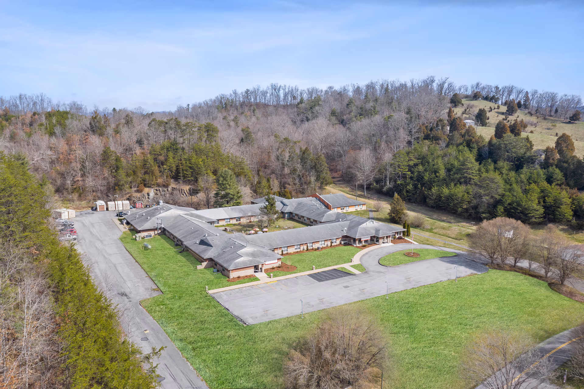 Aerial view of a single-story senior living facility complex with a circular driveway, parking lot and surrounding wooded hills.