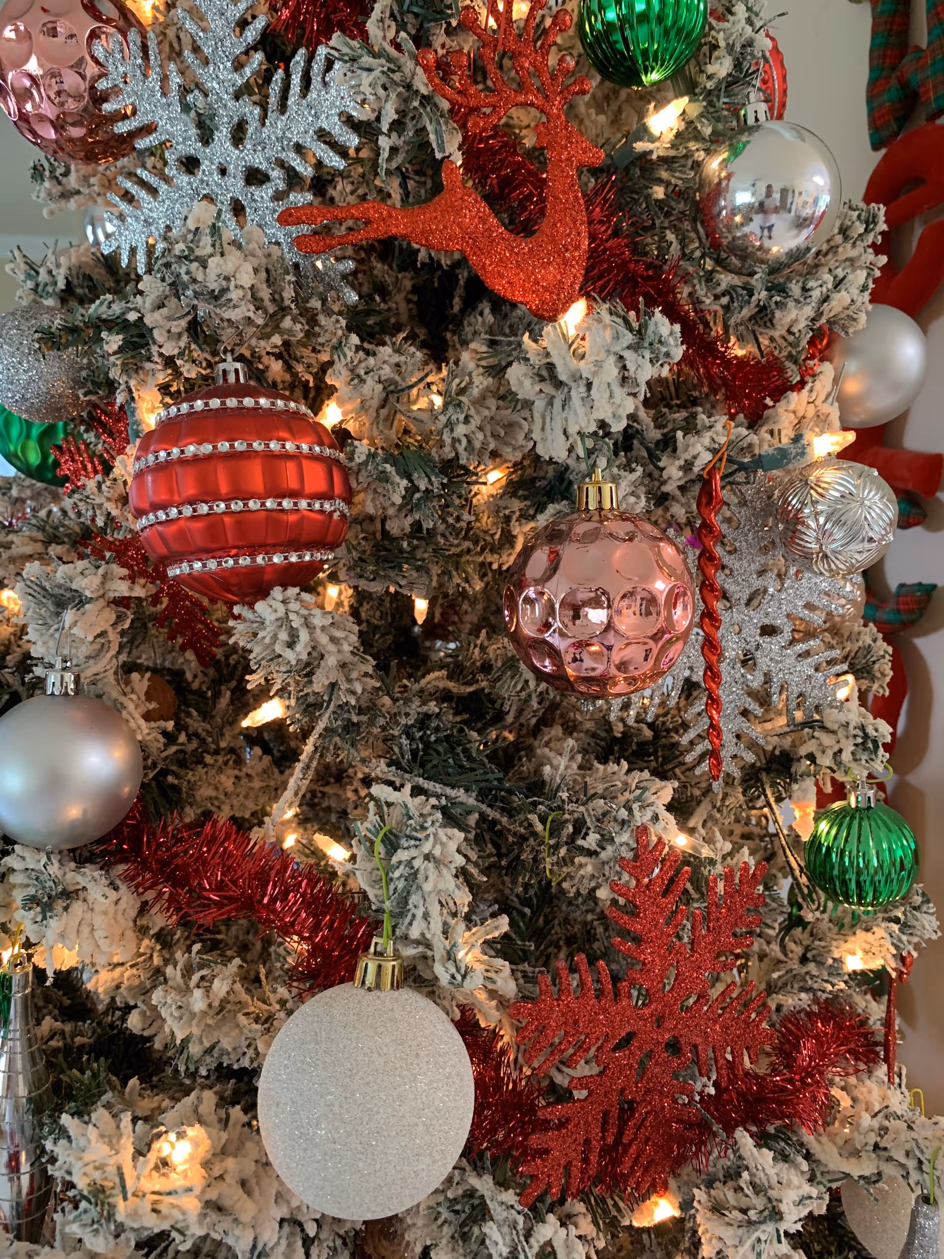 Close-up of a snow-frosted Christmas tree decorated with red, green, silver, and pink ornaments and string lights.