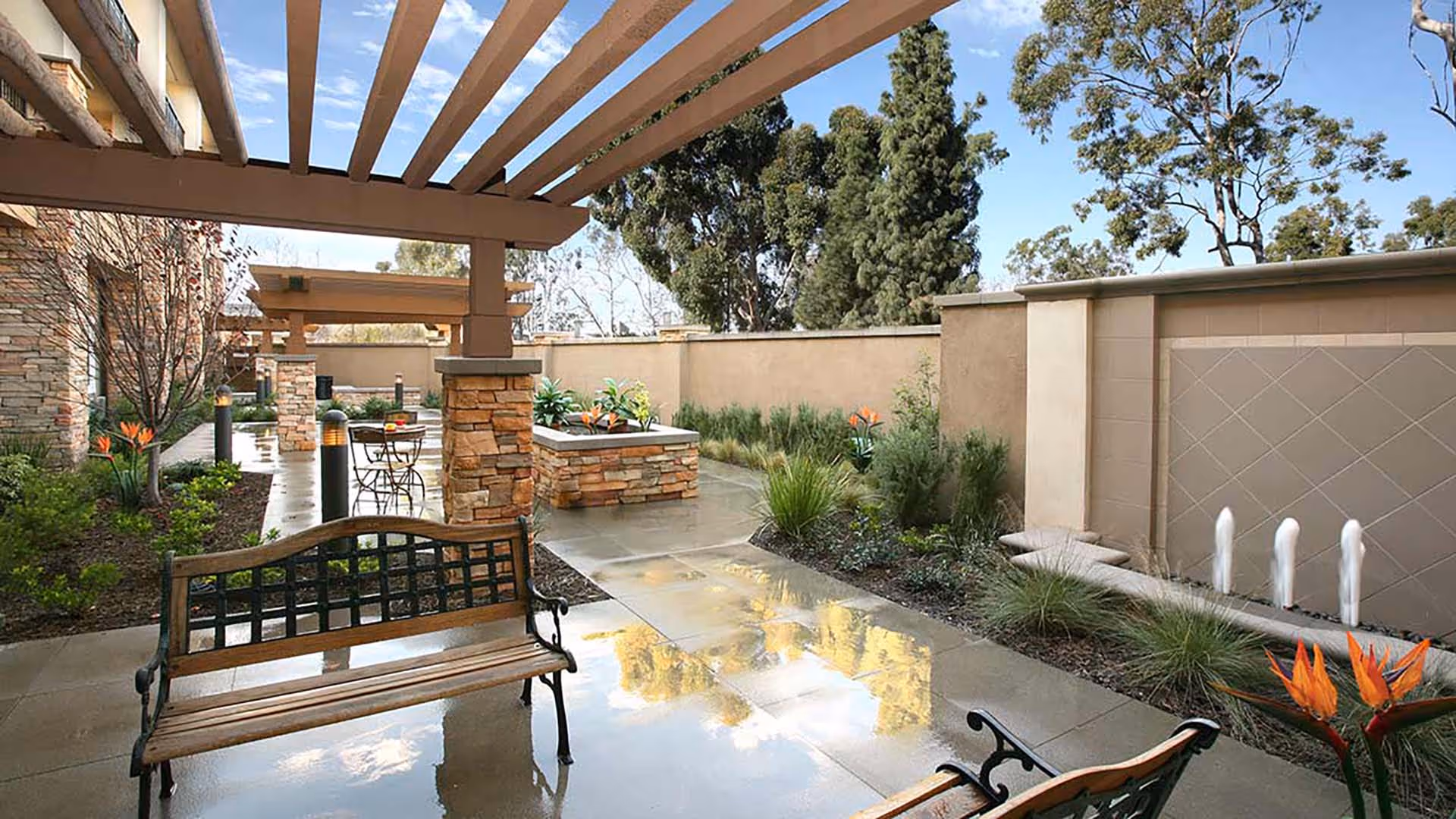Outdoor patio area with wooden benches, stone pillars, a small water fountain, and landscaped plants under a pergola with a clear blue sky and trees in the background.